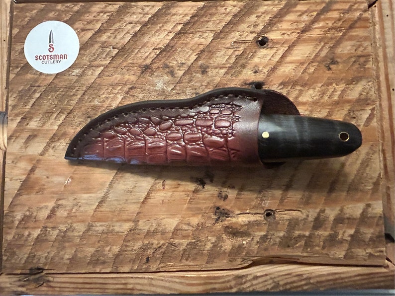 May include: A brown leather knife sheath with a crocodile pattern, a black and brown knife with a gold accent, and the text "Scotsman Cutlery" on a white circle.