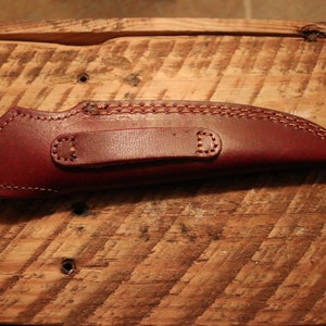 May include: A brown leather knife sheath with a belt loop. The sheath has a stitched design and is on a wooden surface.
