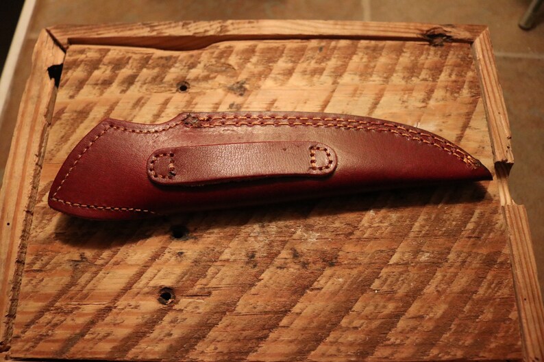 May include: A brown leather knife sheath with a belt loop. The sheath has a stitched design and is on a wooden surface.