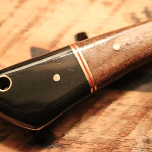 May include: A black and brown knife handle with gold accents. The handle has a hole for a lanyard and a smooth, polished finish.