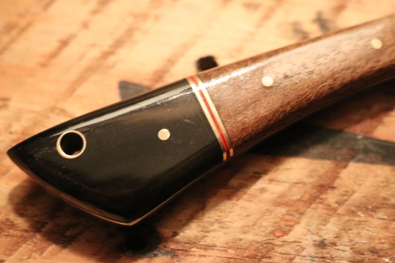May include: A black and brown knife handle with gold accents. The handle has a hole for a lanyard and a smooth, polished finish.