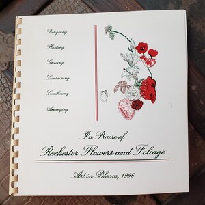 May include: A white booklet with a red and green floral illustration on the cover. The title is "In Praise of Rochester Flowers and Foliage" with the subtitle "Art in Bloom, 1996".