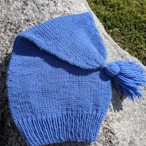 May include: A knitted blue hat with a tassel. The hat is made of a soft, textured yarn and features a folded brim and a tassel at the back. The hat is a solid blue color and is likely handmade.