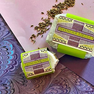 May include: Two bars of lavender-scented soap wrapped in white paper with green and gold labels. The labels read "Savon Surfin" and "Lavender Medley".