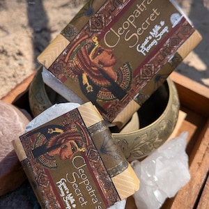 May include: Two bars of soap, wrapped in brown paper, are labelled "Cleopatra's Secret Goat's Milk & Honey Soap." The packaging features an illustration of Cleopatra. The soaps are displayed in a wooden tray with other items.