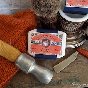 May include: A vintage-style shaving soap bar with the text "Shaving Barbers Favourite Soap Living in the Past" on the label. The soap is on a wooden surface with a shaving brush, a straight razor, and a bowl.