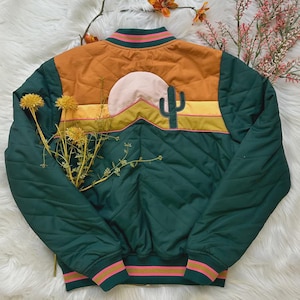 Può includere: Giacca bomber trapuntata nei toni del verde, arancione e giallo. La giacca presenta un design a paesaggio desertico con un cactus, un sole e montagne. I polsini e il colletto hanno strisce rosa e verdi. La giacca è esposta su una superficie bianca e soffice.