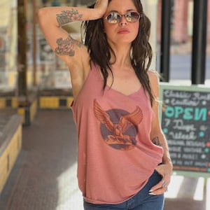 Puede incluir: Camiseta sin mangas rosa polvoriento con un gr&aacute;fico de una criatura alada sobre un fondo circular, combinada con vaqueros azules. Se ve un letrero que dice "Classic Rock Couture OPEN 7 Days a Week".