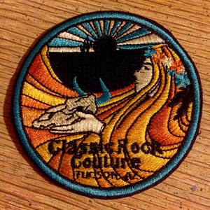 May include: A circular embroidered patch with a blue border and a yellow, orange, and white design. The patch features a stylized image of a woman with flowing hair and a sunburst in the background. The text "Classic Rock Soulvine Tucson, AZ" is embroidered in the lower portion of the patch.