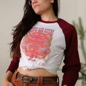 May include: A white baseball-style t-shirt with maroon three-quarter length sleeves. The front features a graphic print of a desert landscape with a scorpion and the text "Desert Rock Couture" and "Arizona". The shirt is tied at the waist.