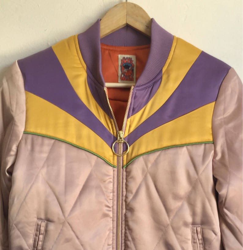 May include: A purple, yellow, and green striped bomber jacket with a quilted design. The jacket has a zipper closure and a small label with the text "Rock and Roll Cowgirl" on the front.