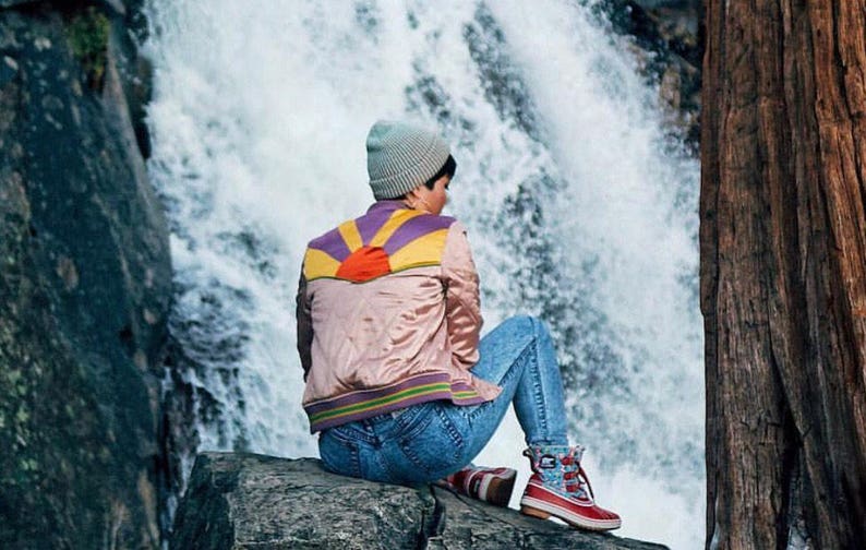 May include: A person wearing a pink bomber jacket with a colorful sunset design on the back, blue jeans, and a light blue beanie sits on a rock in front of a waterfall.