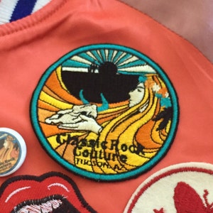 May include: A close-up of a colorful embroidered patch with a woman's face and a sun rising over a mountain. The patch is circular and has a turquoise border. The text "Classic Rock Culture Tucson, AZ" is embroidered in yellow on the patch.
