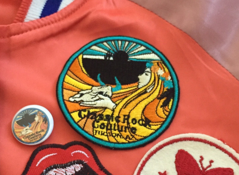 May include: A close-up of a colorful embroidered patch with a woman's face and a sun rising over a mountain. The patch is circular and has a turquoise border. The text "Classic Rock Culture Tucson, AZ" is embroidered in yellow on the patch.