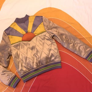 May include: A quilted satin bomber jacket with a colorful sunburst design on the back. The jacket has a purple, yellow, and orange sunburst design with a green and purple trim.