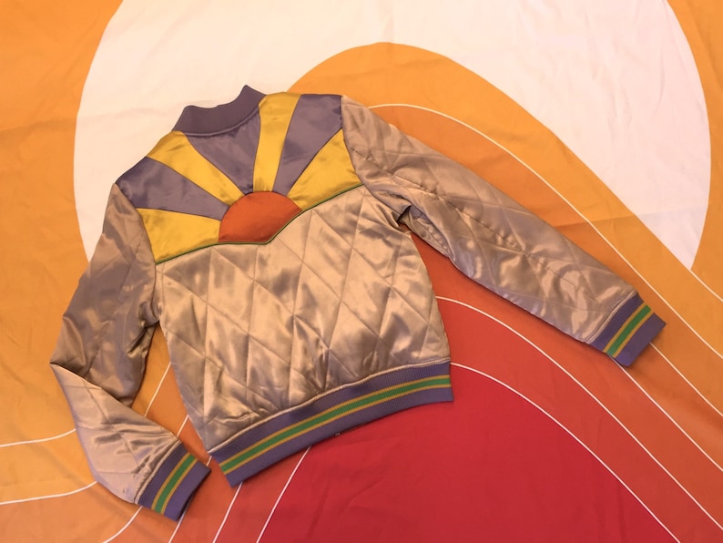 May include: A quilted satin bomber jacket with a colorful sunburst design on the back. The jacket has a purple, yellow, and orange sunburst design with a green and purple trim.