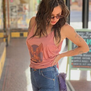 Puede incluir: Una camiseta sin mangas rosa polvoriento con un gr&aacute;fico de un &aacute;guila alada, combinada con vaqueros azules. Un pa&ntilde;uelo morado est&aacute; metido en el bolsillo trasero. El atuendo se usa al aire libre con luz natural.