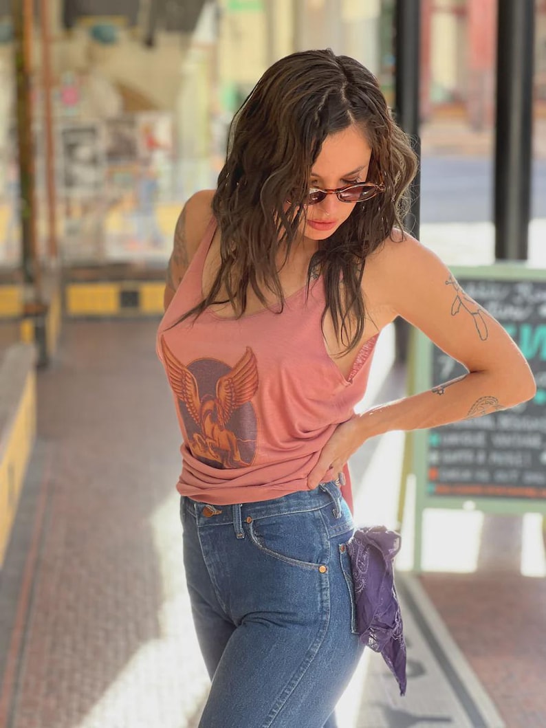 Puede incluir: Una camiseta sin mangas rosa polvoriento con un gr&aacute;fico de un &aacute;guila alada, combinada con vaqueros azules. Un pa&ntilde;uelo morado est&aacute; metido en el bolsillo trasero. El atuendo se usa al aire libre con luz natural.