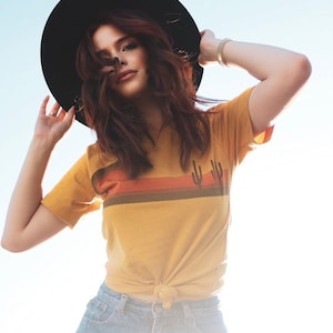 Peut inclure: Une femme portant un t-shirt jaune avec un motif de rayures horizontales et un chapeau noir. Le t-shirt a un motif de cactus sur le devant. Elle porte un short en jean bleu.