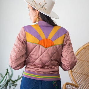 May include: A pink quilted bomber jacket with a colorful sunrise design on the back. The jacket has a green and yellow striped waistband and a white cowboy hat with floral embroidery is worn on top.