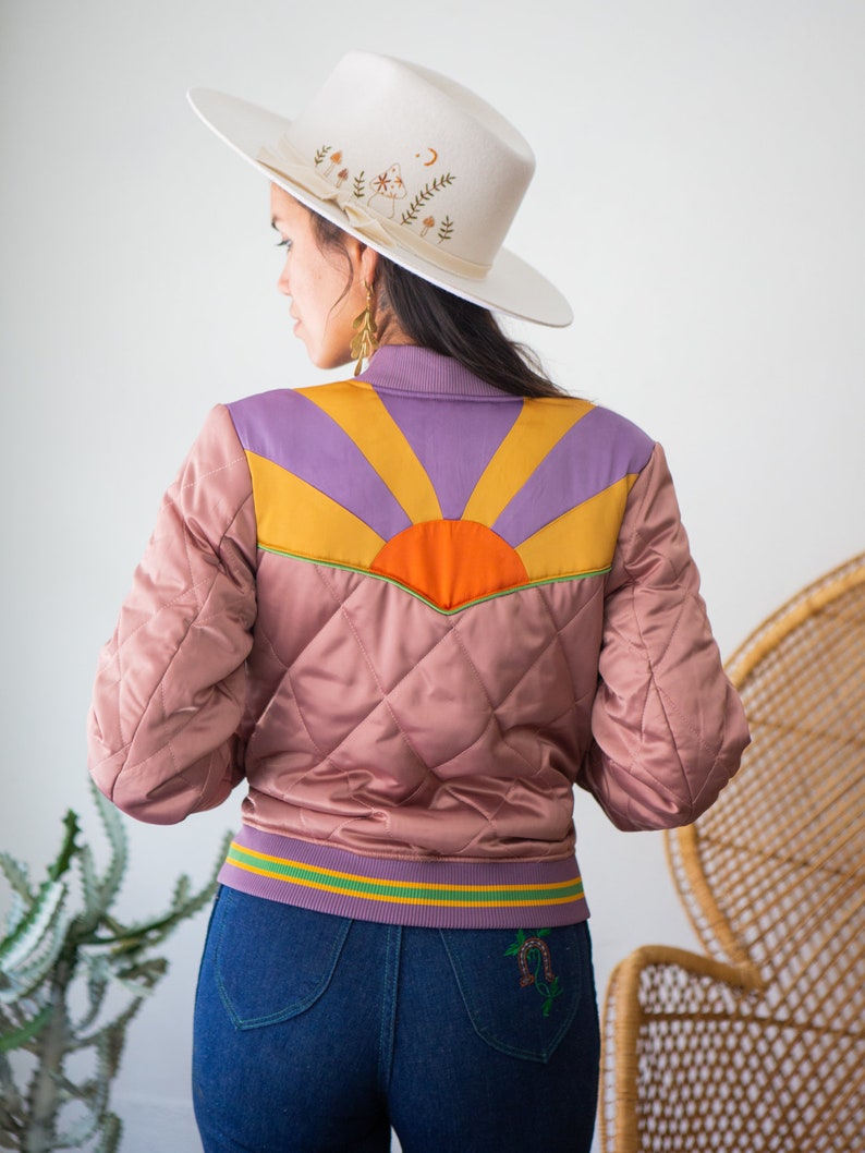 May include: A pink quilted bomber jacket with a colorful sunrise design on the back. The jacket has a green and yellow striped waistband and a white cowboy hat with floral embroidery is worn on top.
