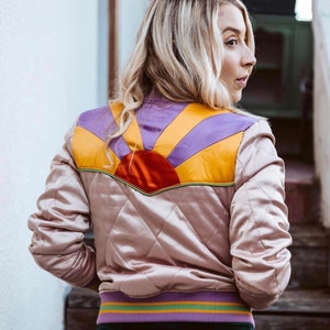May include: A quilted satin bomber jacket in a light pink color with a colorful sunrise design on the back. The design features a red sun with yellow, orange, and purple rays.