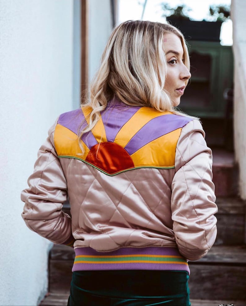 May include: A quilted satin bomber jacket in a light pink color with a colorful sunrise design on the back. The design features a red sun with yellow, orange, and purple rays.