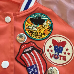May include: A red jacket with various patches, including a tongue with an American flag design, a butterfly with the word "VOTE" and stars, a patch with the words "Classic Rock Culture" and a sun, and a small round patch with a picture of a person.