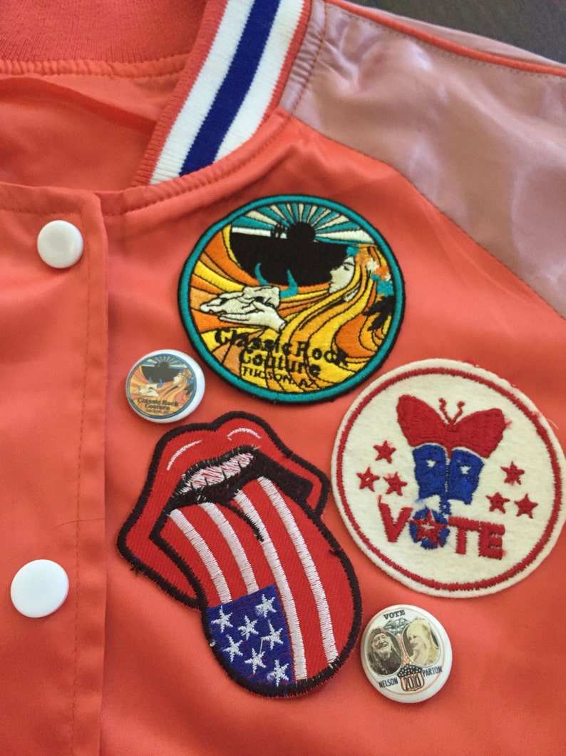 May include: A red jacket with various patches, including a tongue with an American flag design, a butterfly with the word "VOTE" and stars, a patch with the words "Classic Rock Culture" and a sun, and a small round patch with a picture of a person.