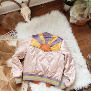 May include: A pink quilted satin bomber jacket with a colorful sunrise design on the back. The jacket has a purple, yellow, and orange sunrise design with a brown outline. The jacket has a green and purple striped trim around the cuffs and bottom.