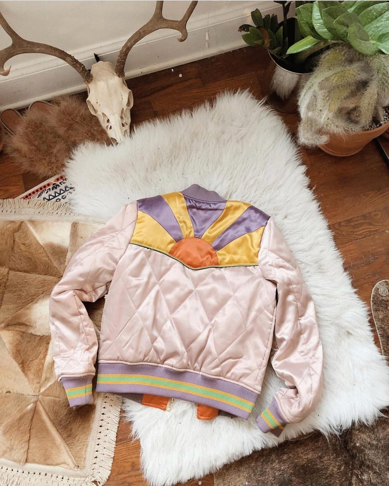 May include: A pink quilted satin bomber jacket with a colorful sunrise design on the back. The jacket has a purple, yellow, and orange sunrise design with a brown outline. The jacket has a green and purple striped trim around the cuffs and bottom.