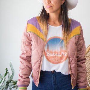 May include: A woman wearing a pink quilted bomber jacket with purple, yellow, and green accents. The jacket has a gold zipper and a white t-shirt underneath with a graphic of a sunset and the text "Bisbee, AZ". She is also wearing blue jeans and a white cowboy hat.