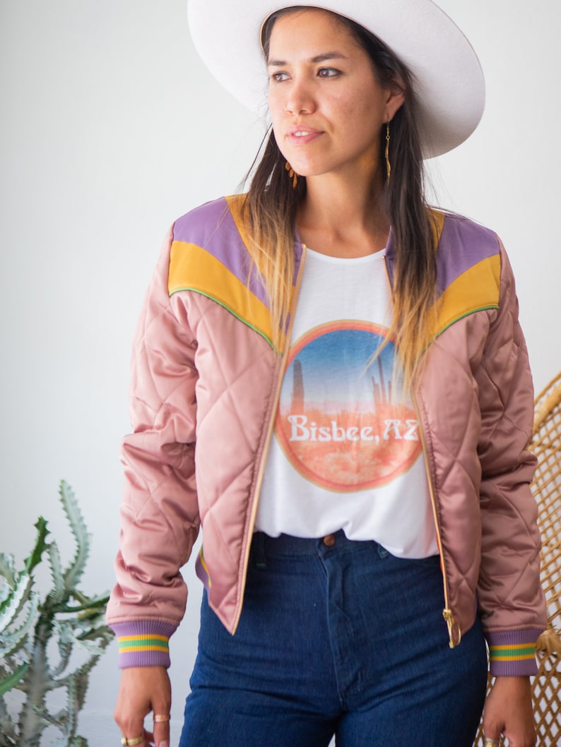 May include: A woman wearing a pink quilted bomber jacket with purple, yellow, and green accents. The jacket has a gold zipper and a white t-shirt underneath with a graphic of a sunset and the text "Bisbee, AZ". She is also wearing blue jeans and a white cowboy hat.