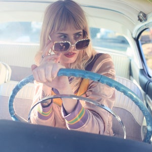 May include: A woman wearing sunglasses and a pink jacket sits in the driver's seat of a vintage car. She is holding the steering wheel with one hand and adjusting her sunglasses with the other.