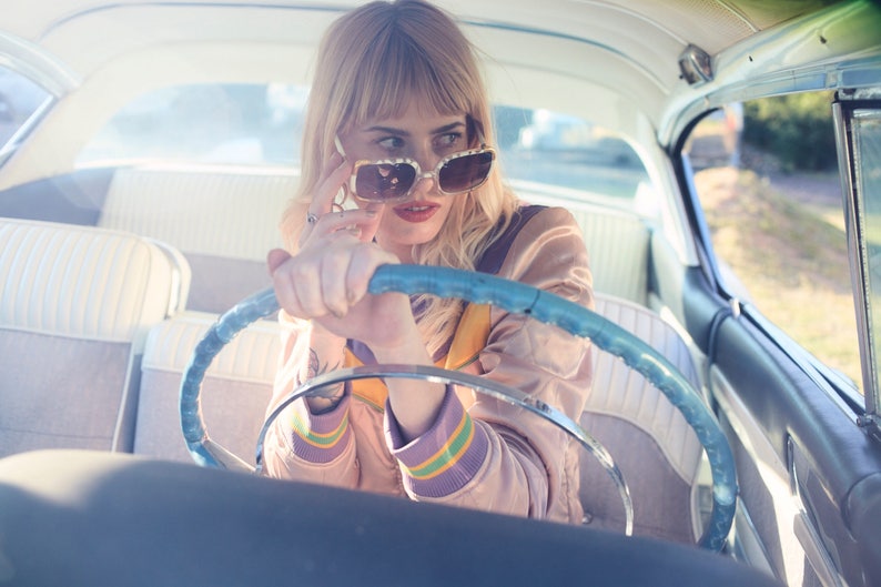 May include: A woman wearing sunglasses and a pink jacket sits in the driver's seat of a vintage car. She is holding the steering wheel with one hand and adjusting her sunglasses with the other.