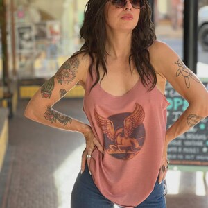 Puede incluir: Una camiseta sin mangas rosa polvoriento con un gr&aacute;fico de un caballo alado dentro de un c&iacute;rculo. La camiseta tiene cuello redondo y se combina con vaqueros azules. La persona lleva gafas de sol y tiene tatuajes en los brazos.