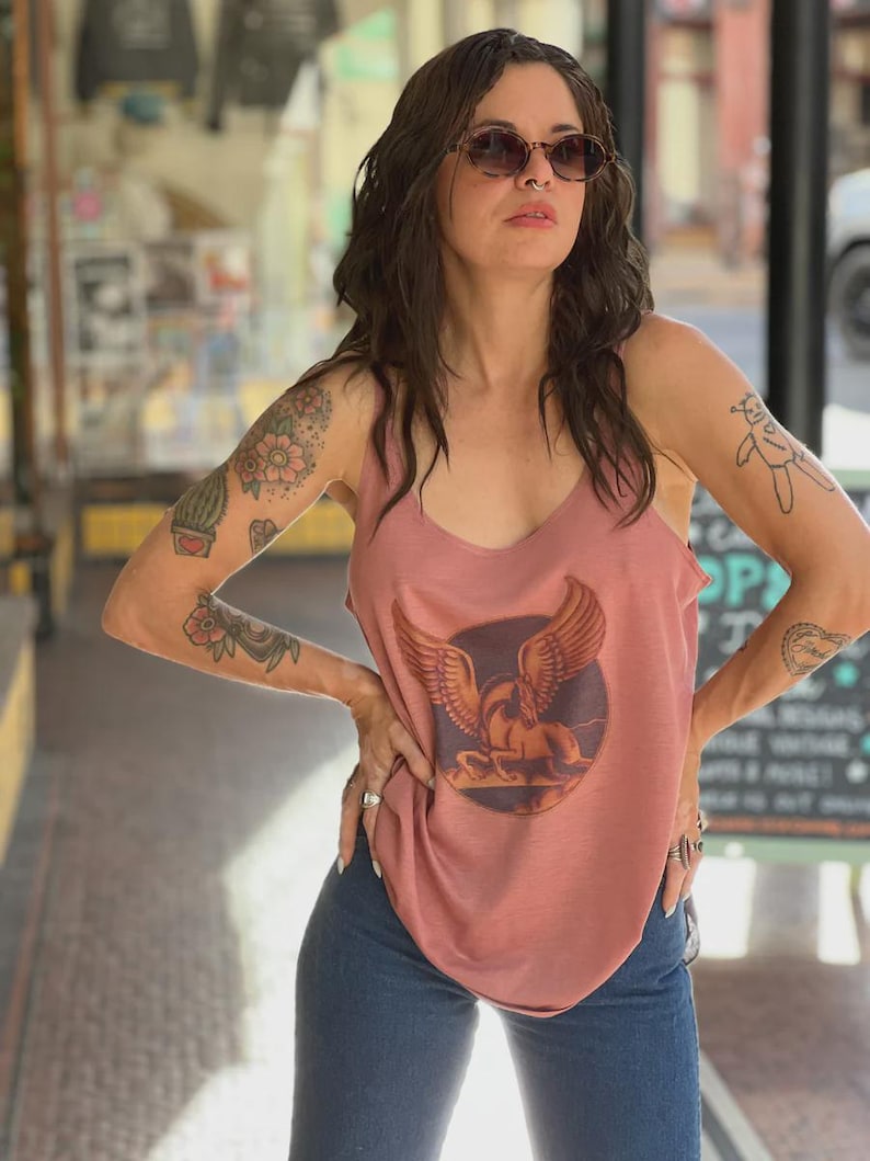 Puede incluir: Una camiseta sin mangas rosa polvoriento con un gr&aacute;fico de un caballo alado dentro de un c&iacute;rculo. La camiseta tiene cuello redondo y se combina con vaqueros azules. La persona lleva gafas de sol y tiene tatuajes en los brazos.