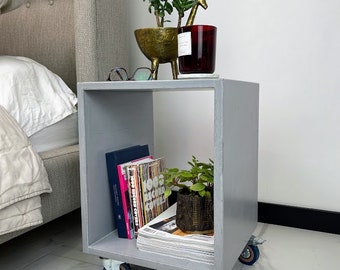 May include: A gray wooden bedside table with wheels, featuring two shelves. The top shelf has a small potted plant, a red candle, and a gold giraffe figurine. The bottom shelf has a stack of books and magazines, and a potted plant.