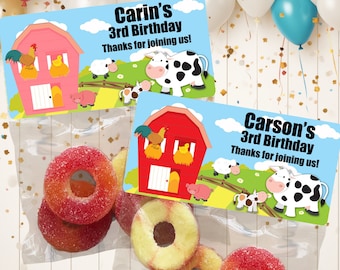 May include: Two clear plastic bags with red and pink farm-themed favor tags. The tags have a red barn, a cow, a pig, and a sheep. The text on the tags reads "Carin's 3rd Birthday Thanks for joining us!" and "Carson's 3rd Birthday Thanks for joining us!" The bags contain red and yellow ring-shaped candies.