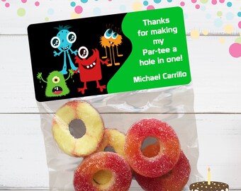 May include: A green and black favor bag with a monster theme. The bag has a sticker that says "Thanks for making my Par-tee a hole in one! Michael Carrillo". The bag contains red and yellow ring-shaped candies.