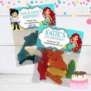May include: Two favor bags with mermaid and prince themed birthday party labels. The bags are filled with colorful gummy candies shaped like fish and dolphins. One label says "Jake & Katie's Birthday!" and the other says "Katie's 4th Birthday!"