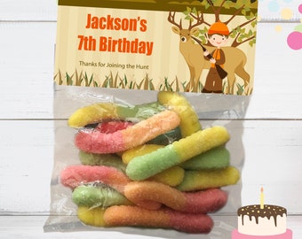 以下が含まれることがあります： カラフルなグミワームが入った透明なビニール袋。袋には「Jackson's 7th Birthday」と書かれたラベルがあり、狩猟をテーマにしたイラストが描かれています。右下には、ろうそくが灯った小さなバースデーケーキがあります。