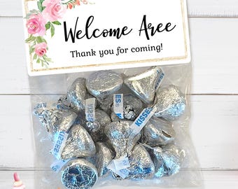 May include: A white favor bag with a gold border and a pink floral design. The bag has a label that says "Welcome Aree" and "Thank you for coming!" The bag is filled with silver foil-wrapped chocolate kisses.