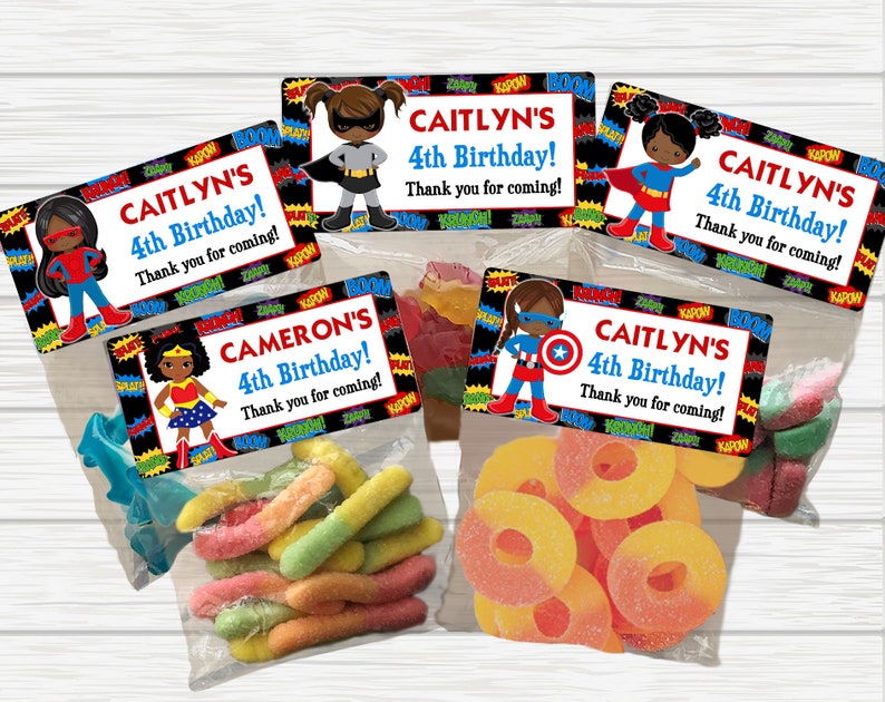 May include: Four favor bags with colorful candy and superhero themed labels. The labels have a black background with colorful text and images of girls dressed as superheroes. The text on the labels reads "CAITLYN'S 4th Birthday! Thank you for coming!" and "CAMERON'S 4th Birthday! Thank you for coming!"