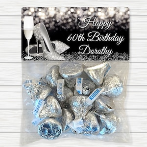 May include: A silver and black favor bag with a label that reads "Happy 60th Birthday Dorothy". The label features a silver high heel shoe and a champagne flute. The bag is filled with silver foil-wrapped Hershey's Kisses.