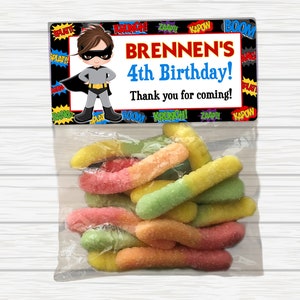 May include: A superhero-themed party favor bag with a colorful comic book design. The bag contains a variety of colorful gummy worms. The bag has a label that says "BRENNEN'S 4th Birthday! Thank you for coming!"