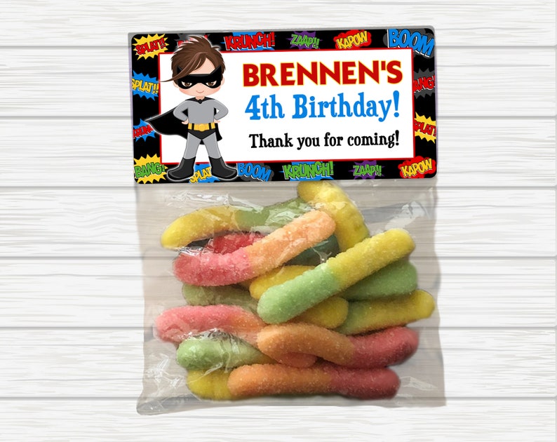May include: A superhero-themed party favor bag with a colorful comic book design. The bag contains a variety of colorful gummy worms. The bag has a label that says "BRENNEN'S 4th Birthday! Thank you for coming!"
