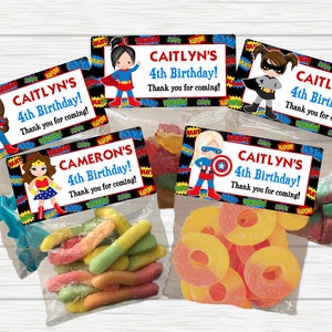 May include: Four colorful party favor bags with superhero themed labels. The labels feature a girl dressed as a superhero with the text "CAITLYN'S 4th Birthday! Thank you for coming!" The bags contain colorful gummy candies.