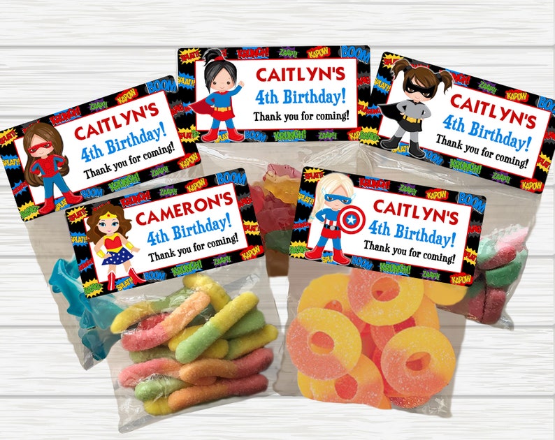 May include: Four colorful party favor bags with superhero themed labels. The labels feature a girl dressed as a superhero with the text "CAITLYN'S 4th Birthday! Thank you for coming!" The bags contain colorful gummy candies.