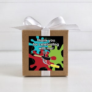 May include: A small brown box with a white ribbon tied around it. The box has a square sticker on the front with a black background, colourful paint splatters, a paintball gun, and the text "Thank you for celebrating with me!"
